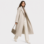 A woman models a long, light beige wool coat over a taupe turtleneck, cream wide-leg trousers, and beige heeled boots, while carrying a taupe leather shoulder bag against a solid light gray background.