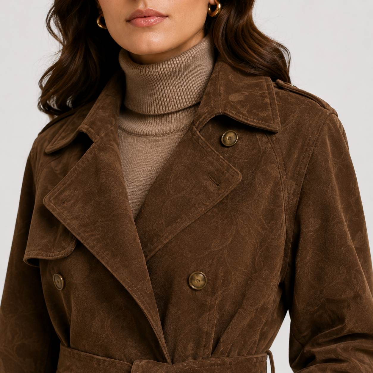 A close-up of a model wearing a brown textured suede trench coat over a beige ribbed turtleneck sweater, set against a plain light gray background.