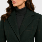 A close-up of a woman wearing a dark green windowpane plaid blazer over a dark charcoal turtleneck sweater, with a small gold hoop earring visible.