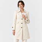 A woman modeling a light beige trench coat with brown buttons, worn over a white top and white trousers, against a plain white studio background.