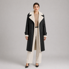 A model stands against a light gray studio background wearing a long black faux-leather trench coat with a thick cream shearling-style collar and lining, paired with cream trousers, a beige top, and black heels.