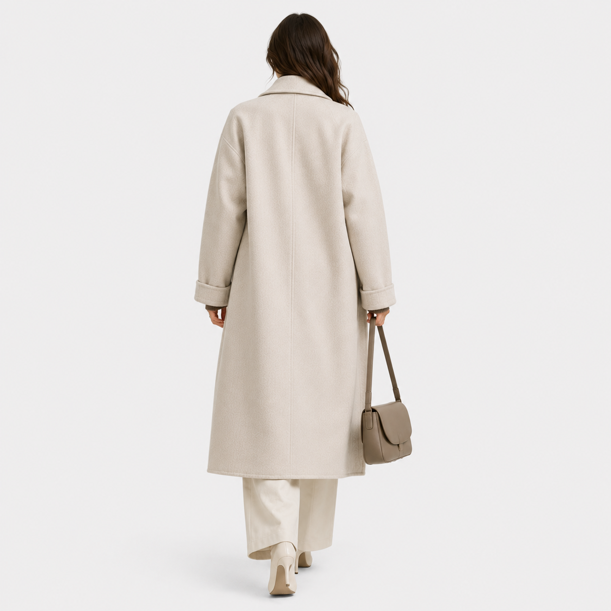 A woman shown from behind wearing a long cream-colored coat, matching wide-leg trousers, and beige heels, while carrying a taupe shoulder bag against a light gray studio background.