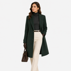 A woman models a dark green windowpane plaid coat over a black turtleneck and cream trousers, holding a brown handbag against a plain white studio background.