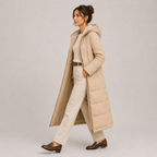 A woman models a long, light beige hooded puffer coat over a cream top and cream trousers, paired with brown loafers, against a neutral light gray studio background.