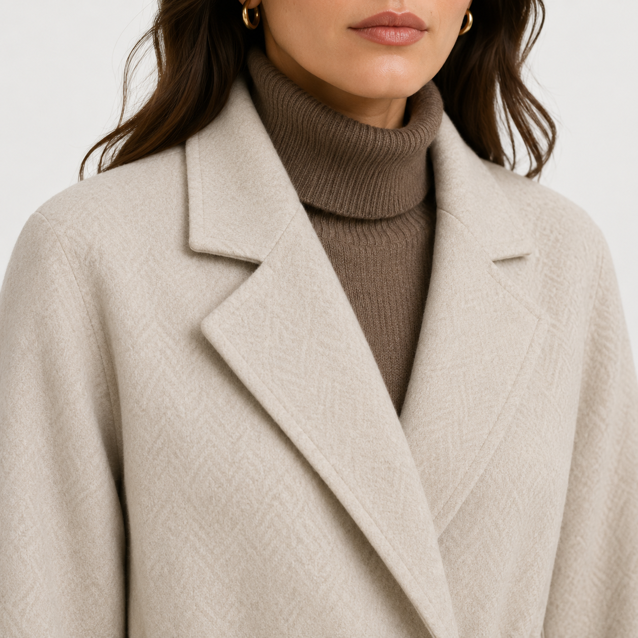 A close-up of a woman wearing a light beige textured coat over a brown ribbed turtleneck sweater, against a clean light gray background.