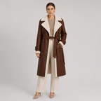 A woman models a long brown faux-leather trench coat with a cream-colored shearling-style collar and lining, styled with cream pants, a beige top, and beige heels against a light gray studio background.