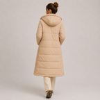 A woman viewed from behind wearing a long, light beige hooded puffer coat over cream pants and brown shoes, standing against a plain light gray studio background.