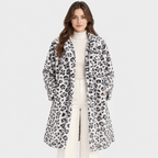 A woman modeling a long white faux-fur coat with a black leopard print pattern, worn over a cream turtleneck and cream pants, against a light gray studio background.