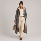 A woman models a long gray puffer coat with a beige sherpa lining, styled with cream trousers, a cream top, a brown belt, brown loafers, and a taupe handbag against a light gray studio background.