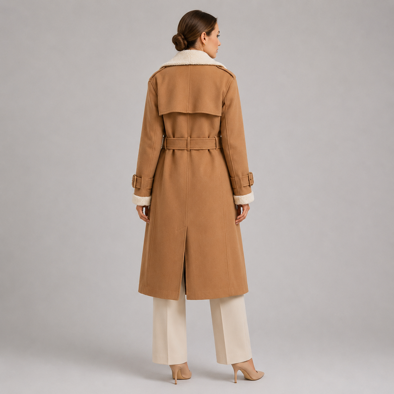 A woman shown from the back wearing a camel-colored trench coat with a cream shearling-style collar and cuffs, paired with cream wide-leg pants and beige heels, against a neutral gray studio background.