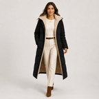A woman models a long black puffer coat with a beige sherpa-lined hood and interior, worn over a cream sweater, cream pants, and brown ankle boots, against a light gray studio background.