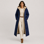 A woman models a long navy blue puffer coat with a beige sherpa-lined hood and interior, styled over a cream top and trousers with a brown belt and boots, against a light gray studio background.