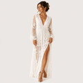 A woman modeling a long white lace maxi dress with sheer long sleeves, a deep V-neckline, and a high side slit, photographed against a plain light gray studio background.