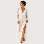 A woman modeling a long white lace maxi dress with sheer long sleeves, a deep V-neckline, and a high side slit, photographed against a plain light gray studio background.