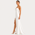 A woman modeling a floor-length white satin gown with thin straps, a ruched bodice, and a high front slit, photographed against a plain light gray studio background.