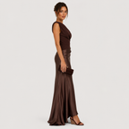 A woman modeling a floor-length, sleeveless dark brown gown with a draped neckline and a satin-like skirt, standing against a plain light gray studio background.