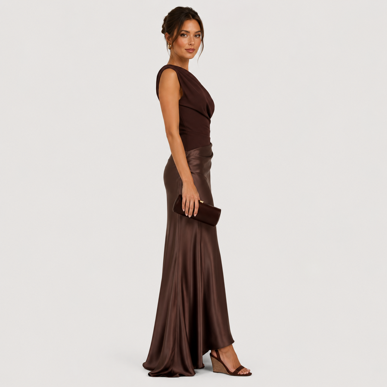 A woman modeling a floor-length, sleeveless dark brown gown with a draped neckline and a satin-like skirt, standing against a plain light gray studio background.