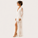 A woman modeling a long white lace maxi dress with sheer long sleeves, a deep V-neckline, and a high thigh slit, shown against a plain light background.