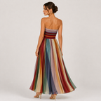 A woman stands with her back to the camera wearing a strapless maxi dress with a multicolor striped tulle skirt and a striped bodice, set against a plain light gray studio background.