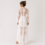 A woman viewed from the back wearing a long white lace maxi dress with sheer panels, floral embroidery, and a high neckline, photographed against a plain light gray studio background.