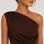 A close-up of a model wearing a dark brown, one-shoulder draped top against a light gray background.