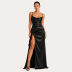 A woman modeling a floor-length black satin gown with thin straps, a ruched bodice, and a high thigh slit, photographed against a plain light gray studio background.