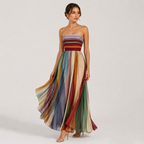 A woman models a strapless maxi dress with a multicolor striped bodice and flowing pleated skirt in shades of blue, green, yellow, orange, and red, set against a plain light gray studio background.