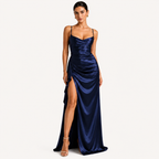 A woman modeling a floor-length navy satin gown with thin straps, a draped bodice, and a high slit, photographed against a plain light background.