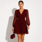 A woman models a short burgundy dress with a V-neckline, long sheer puff sleeves, and a flared skirt with subtle glittery speckles. She is holding a matching burgundy handbag with a gold chain strap against a plain white background.