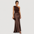 A woman models a floor-length one-shoulder satin gown in a deep chocolate brown color, paired with a matching dark brown clutch and brown heeled sandals, set against a solid light gray background.