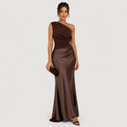A woman models a floor-length one-shoulder satin gown in a deep chocolate brown color, paired with a matching dark brown clutch and brown heeled sandals, set against a solid light gray background.