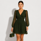 A woman modeling a dark green sparkly mini dress with sheer long sleeves and a V-neckline, holding a matching green handbag against a plain light gray background.