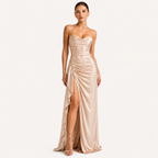 A woman modeling a floor-length champagne satin gown with thin straps, a ruched bodice, and a high draped slit, photographed against a plain light gray studio background.
