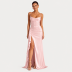 A woman modeling a floor-length light pink satin gown with thin spaghetti straps, a sweetheart neckline, ruched detailing, and a high side slit, photographed against a plain light gray studio background.