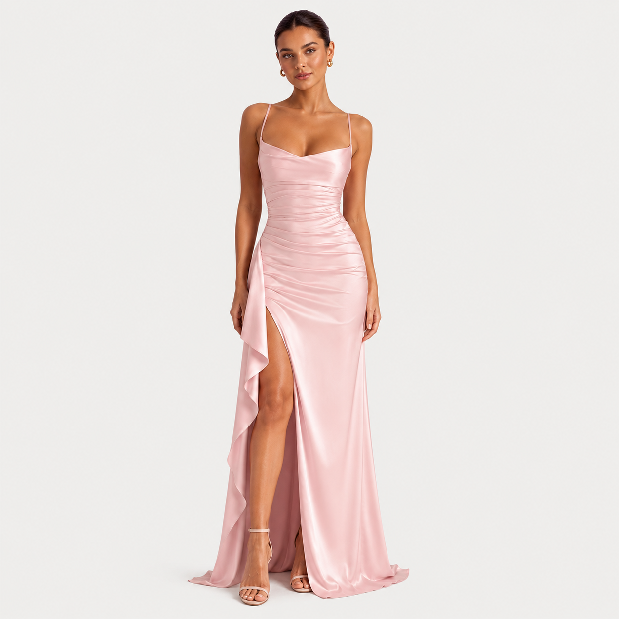 A woman modeling a floor-length light pink satin gown with thin spaghetti straps, a sweetheart neckline, ruched detailing, and a high side slit, photographed against a plain light gray studio background.