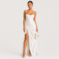 A woman modeling a white satin slip dress with thin straps, a draped ruched bodice, and a high thigh slit, standing against a light gray studio background and holding a small cream handbag.