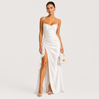 A woman modeling a white satin slip dress with thin straps, a draped ruched bodice, and a high thigh slit, standing against a light gray studio background and holding a small cream handbag.