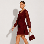 A woman models a sparkling burgundy mini dress with sheer long balloon sleeves and a V-neckline, holding a matching burgundy handbag with a gold chain strap against a light gray studio background.