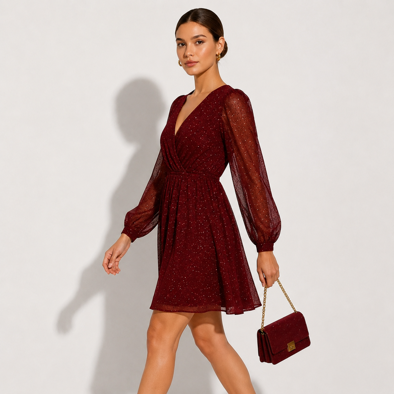 A woman models a sparkling burgundy mini dress with sheer long balloon sleeves and a V-neckline, holding a matching burgundy handbag with a gold chain strap against a light gray studio background.