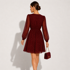 A woman stands with her back to the camera wearing a short burgundy dress with sheer long sleeves and a flared skirt. She is holding a matching burgundy handbag against a plain light gray studio background.