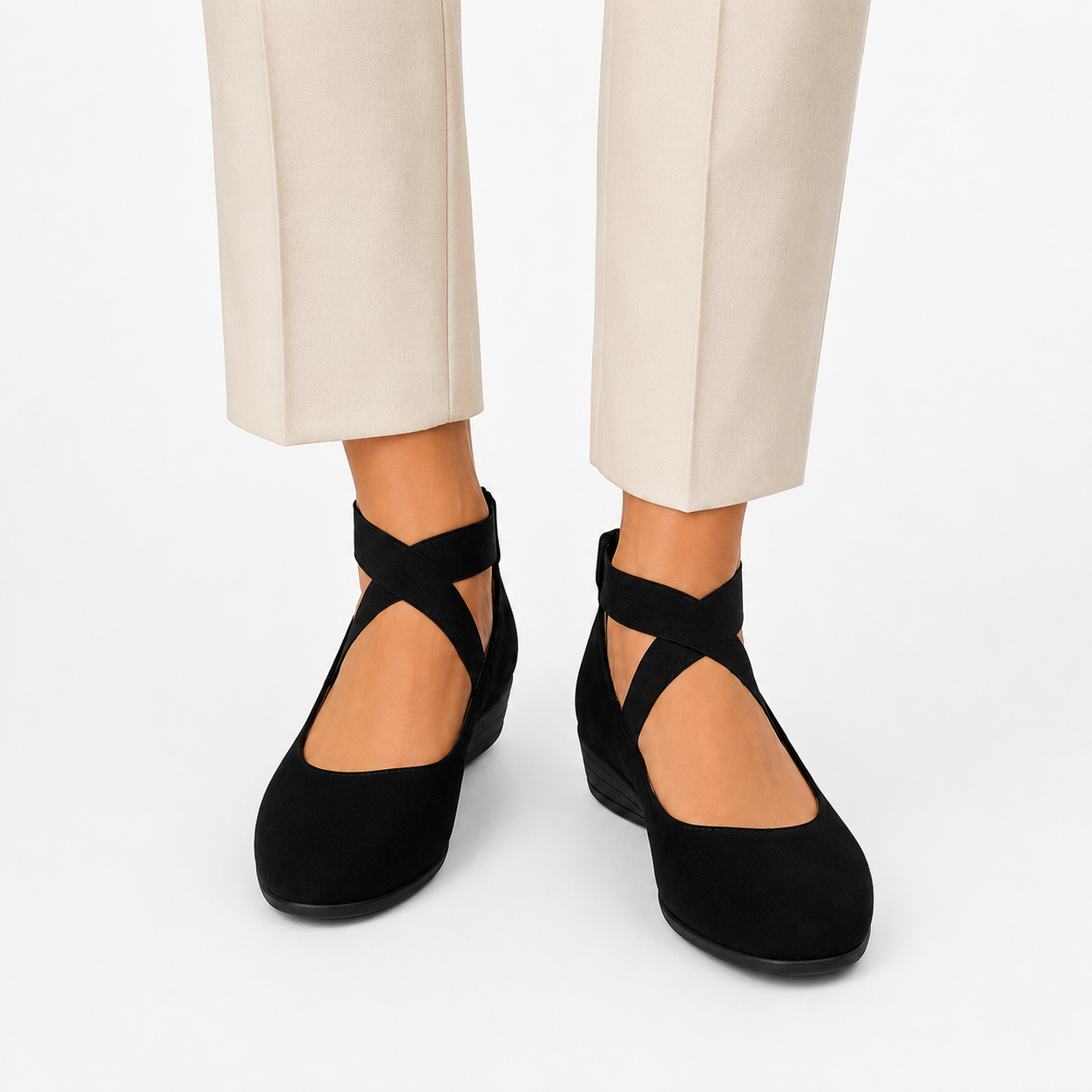 Black flat shoes with crisscross ankle straps worn by a model in beige trousers, shown against a plain white studio background.