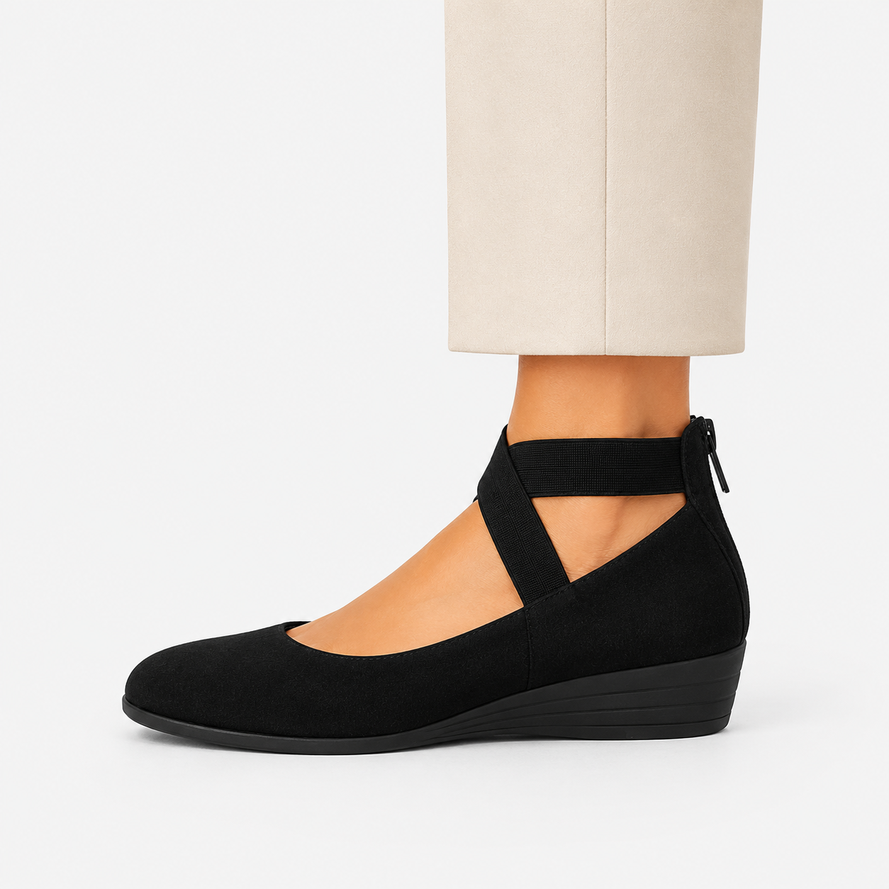 A black wedge shoe with an ankle strap and cutout details, shown on a model's foot with beige trousers against a light gray background.