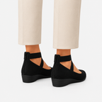 Black wedge shoes with ankle straps worn with cream-colored trousers, shown from a rear three-quarter angle against a light gray background.