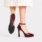 A pair of glossy deep burgundy patent leather high-heeled pumps with ankle straps and gold buckle details, shown on a model's feet against a light gray background.