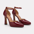 A pair of glossy burgundy patent leather high-heeled pumps with ankle straps and gold-tone buckle details, displayed on a clean light gray background.