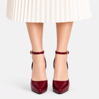 A pair of glossy deep burgundy ankle-strap high heels with gold buckle details, shown on a model's feet with a white pleated skirt visible at the top.