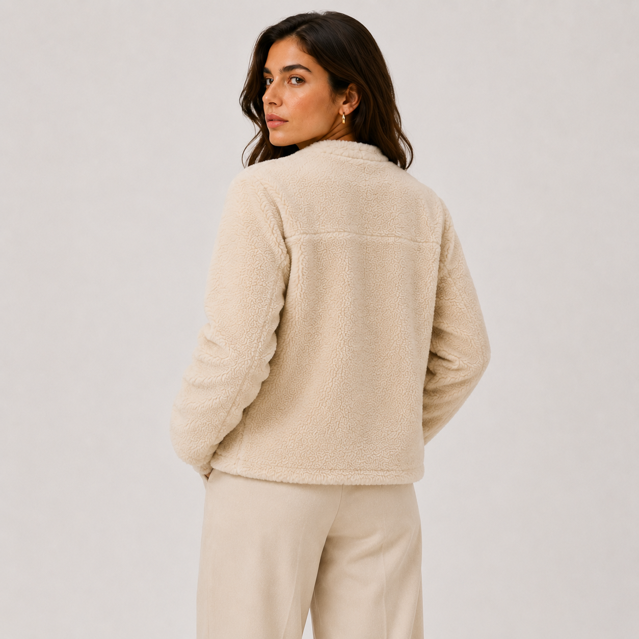A woman shown from the back wearing a cream-colored sherpa fleece jacket and matching cream trousers, photographed against a neutral light gray studio background.