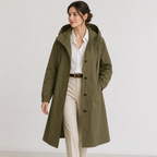A woman wearing an olive green hooded coat over a white button-up shirt and beige trousers, standing against a plain light gray studio background.