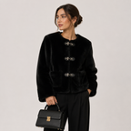 A woman models a black cropped faux-fur jacket with decorative toggle closures, paired with black trousers and holding a black handbag, set against a plain light gray studio background.