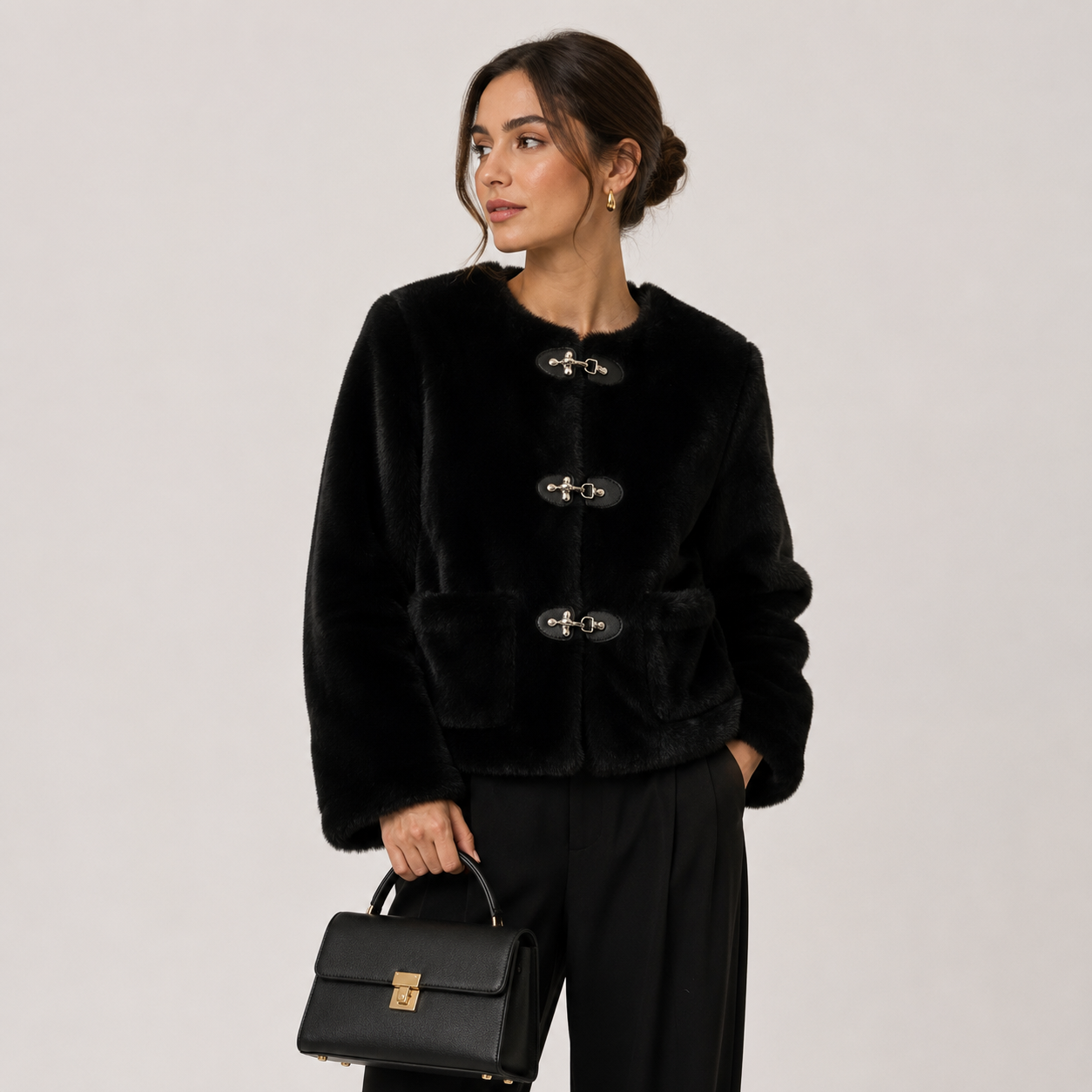 A woman models a black cropped faux-fur jacket with decorative toggle closures, paired with black trousers and holding a black handbag, set against a plain light gray studio background.